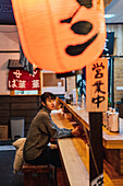 Asiatische Frau in Freizeitkleidung sitzt am Holztresen und wartet auf ihre Bestellung in einer Ramen-Bar