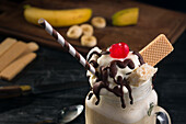 From above of glass jar of sweet banana split milkshake topped with whipped cream waffles chocolate and cherry on table