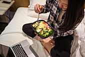 From above Asian female eating healthy salad and watching video on netbook while sitting on bed in light hotel room during quarantine