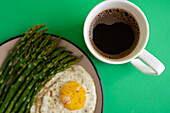 From above breakfast dish of fried egg with green asparagus server on plate next to cup of coffee on yellow background