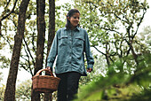 From below female walking through trees and collecting mushrooms in wicker basket in foggy forest