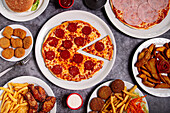Blick von oben auf einen Teller mit einer leckeren Peperoni-Pizza und einer Schüssel mit Soße und verschiedenen Junkfood-Gerichten auf einem grauen Tisch beim Mittagessen