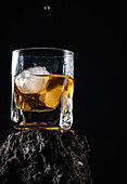 Whiskeytropfen fallen auf Eiswürfel in einem Kristallglas auf einer rauen Oberfläche vor schwarzem Hintergrund