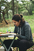 Concentrated female sitting at table and taking notes in notebook in green park in daytime
