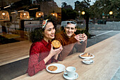 Fröhliches, verliebtes, junges, gemischtrassiges Paar sitzt an einem Holztresen und genießt köstliches Gebäck und Kaffee in einer gemütlichen Cafeteria