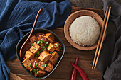 Close up mala tofu, chinese vegan dish, accompanied by a bowl of rice on top of a wooden table decorated with fabrics