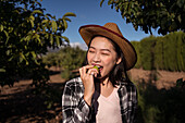 Delighted ethnic female farmer in straw hat and checkered shirt eating fresh tasty apple while standing in orchard in countryside on sunny day