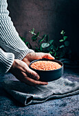 Hand of crop unrecognizable person holding a bowl with raw red lentils on table with towel and green plants in light kitchen