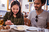 Happy young Asian lady with dark hair in casual clothes smiling while eating delicious salad during lunch with ethnic boyfriend in restaurant