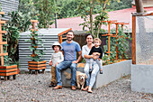 Ganzkörperaufnahme von glücklichen Eltern mit niedlichen Söhnen, die mit einem Golden Retriever Welpen auf einem Gartenbeet sitzen und in die Kamera schauen