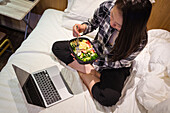 From above Asian female eating healthy salad and watching video on netbook while sitting on bed in light hotel room during quarantine