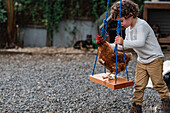 Little curly haired boy swing poultry chicken while playing in garden in village