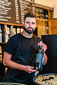 Seriöser bärtiger Barkeeper mit dunklem Haar in schwarzer Schürze steht am Tresen und zeigt eine Flasche Rotwein, während er im Restaurant arbeitet