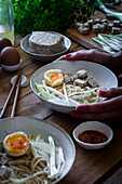 Von oben abgeschnittene, nicht erkennbare Person isst frisch gekochte Ramen-Nudeln mit Tofu, Eiern und Gemüse mit Stäbchen auf einem Holztisch