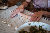 From above of crop anonymous woman in apron rolling dough on table while preparing dumplings with meat in kitchen