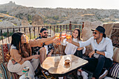 Hochformatige Aufnahme von lächelnden Freunden, die um einen Tisch herum sitzen und ihre Gläser mit Cocktails erheben, während sie in einer Bar auf einer Terrasse in Kappadokien, Türkei, abhängen