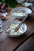 Ausschnitt einer nicht erkennbaren Person, die frisch gekochte Ramen-Nudeln mit Tofu, Eiern und Gemüse mit Stäbchen auf einem Holztisch zubereitet
