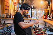 Side view of man in bandana standing at counter and cooking ramen in modern Asian cafe