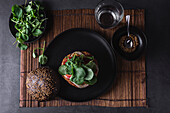 Appetizing burger with sesame seeds placed on black plate near salad leaves against glass of water on dark background