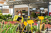 Busy female gardener in mask standing near various plants and browsing tablet while working in garden center
