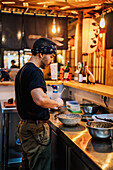 Side view of man in bandana standing at counter and cooking ramen in modern Asian cafe