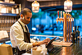 Seriöser männlicher Barkeeper in Uniform und Maske, der während einer Coronavirus-Pandemie an der Theke einer Bar an einem Tablet arbeitet