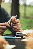 Crop unrecognizable person sitting at table and cutting fresh mushroom against green grass in nature