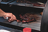 Eine gesichtslose Person mit einer Zange grillt Fleisch auf einem Rost in einem heißen Grill in der Nähe von Soße während des Kochvorgangs