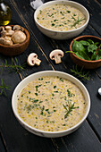 White bowls with tasty fresh homemade mushroom soup served on black table with green herbs in light kitchen at home