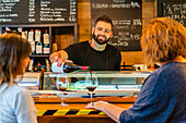Concentrated barman standing at counter and pouring red wine into glasses for unrecognizable woman customer in cafe