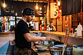 Seitenansicht eines Mannes mit Kopftuch, der an der Theke steht und Ramen in einem modernen asiatischen Café kocht