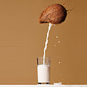 Milk pouring from fresh whole coconut into glass served on table against brown background