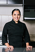 Smiling ethnic female chef in black professional uniform standing at metal table in modern kitchen in restaurant