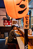 Seitenansicht einer jungen asiatischen Frau in Freizeitkleidung, die an einem Holztresen sitzt und auf ihre Bestellung in einer Ramen-Bar wartet