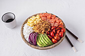 From above bowl of tasty poke dish with raw ingredients placed near chopsticks and cup of soy sauce on white plaster surface