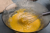 Mehl gießt in eine Glasschüssel mit rohem, aufgeschlagenem Eigelb und Schneebesen, die während des Kochvorgangs auf dem Tisch in der Küche steht