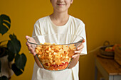 Unkenntlich gemachtes lächelndes Teenager-Mädchen in weißem T-Shirt zeigt eine große Glasschüssel mit gehacktem gesundem Gemüse zu Hause
