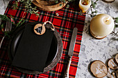 Top view of christmas table setting with empty menu on ceramic plate and red checkered tablecloth with yellow lights on the background