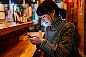 Seitenansicht einer zufriedenen Asiatin im legeren Pullover, die lächelt, während sie am Tresen einer traditionellen Ramen-Bar ihr Mobiltelefon benutzt
