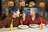 Durch ein Glas lachende ethnische Frau füttert ihren Freund mit köstlichem vegetarischem Salat, während sie gemeinsam eine gesunde Mahlzeit an der Theke eines modernen Restaurants einnimmt