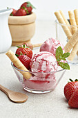 Delicious homemade strawberry ice cream scoops served in glass bowl with berries and waffle roll in kitchen in daylight