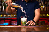 Unrecognizable bartender pouring crushed ice to the glass while preparing mojito cocktail in the bar