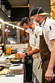 Seitenansicht eines professionellen Kochs mit Maske, der mit einem Kollegen in einer Restaurantküche während einer Coronavirus-Pandemie einen Teller mit einem köstlichen Gericht serviert