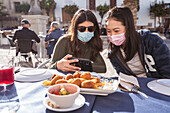 Positive Asian woman with female friend in medical masks watching video on cellphone while sitting at table in outdoor cafeteria