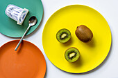 Top view of fresh ripe whole and halved kiwis and yogurt with spoon placed on colorful ceramic plates on white table