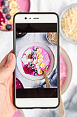 Top view of crop unrecognizable person demonstrating photography of delicious breakfast bowl with smoothie and fruits on cellphone screen