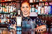 Smiling female barkeeper standing at bar counter with a type of alcohol drink served in creative cocktail glasses in shape of jellyfish