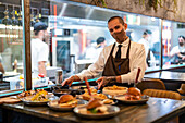 Stilvoller männlicher Kellner in Uniform stellt Teller mit verschiedenen Speisen auf den Tisch in einem Restaurant mit offener Küche während der COVID-Pandemie