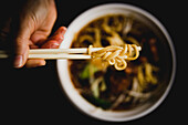 Nahaufnahme der Hand einer anonymen Person, die mit Stäbchen Nudeln aus einer Schüssel mit leckerer asiatischer Suppe nimmt