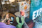 Through window back view of crop unrecognizable male with delicious beverage in takeaway glass using app on cellphone in restaurant